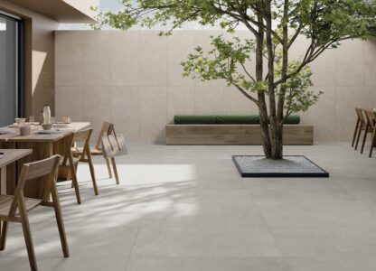 Spazio outdoor. A pavimento, finitura Grey, grigio scuro e dalla forte personalità, nel formato 60x120, spessore 20 mm da esterni. Sulla parete frontale è visibile il decoro Weave (60x120) nella variante Clay, che rievoca le texture Art Decò. La panca è rivestita con la collezione Unica, colore Beige, nel formato 20x120